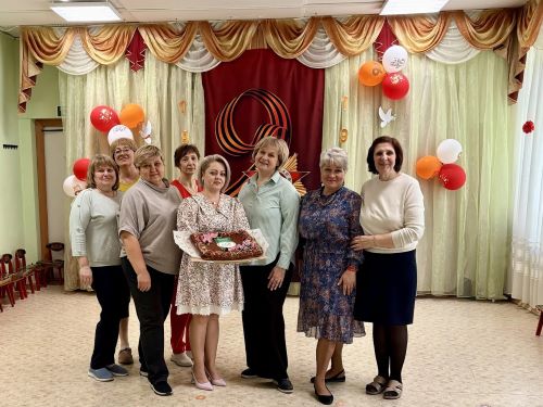 На фотографии изображена группа из семи женщин, которые позируют для совместного снимка в празднично украшенном зале. Женщины стоят в один ряд, улыбаются и выглядят радостными. Одна из них держит в руках большой торт, украшенный декором.  Фон — декорированный зал с драпированными шторами светлых и золотистых оттенков. В центре фона расположен большой бордовый баннер с абстрактным или символическим рисунком. По обе стороны от баннера развешаны воздушные шары красного, белого и оранжевого цветов. На полу — светлое покрытие. Атмосфера снимка праздничная, торжественная.